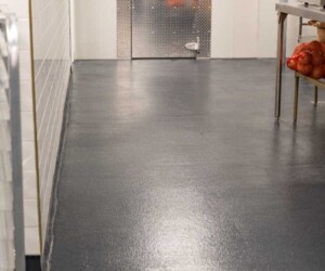 A clean commercial kitchen area with USDA approved flooring, a shiny dark surface, metal prep table holding onions on the right, white tiled wall on the left, and a closed walk-in freezer door at the end of the room. A clean commercial kitchen area with USDA approved flooring, a shiny dark surface, metal prep table holding onions on the right, white tiled wall on the left, and a closed walk-in freezer door at the end of the room.