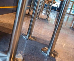 Close-up view of shiny metal poles anchored to polished, speckled commercial kitchen flooring inside a building, with tables and windows visible in the background. Close-up view of shiny metal poles anchored to polished, speckled commercial kitchen flooring inside a building, with tables and windows visible in the background.